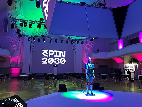 Androidin leerer Saal, bunt beleuchtet, mit Androidin auf Bühne in der Mitte