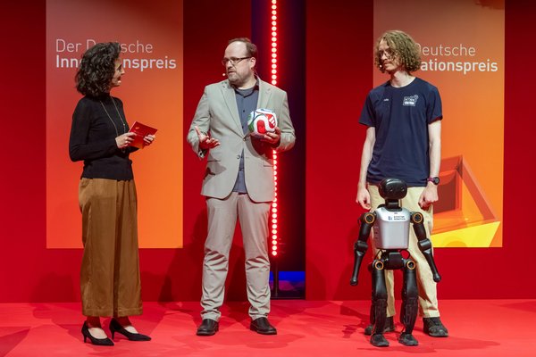 Roboterfußball auf der Bühne des Deutschen Innovationspreis Moderatorin im Gespräch mit Prof. Crönertz, der einen Fußball hält, und Felix Loos mit humanoiden Roboter