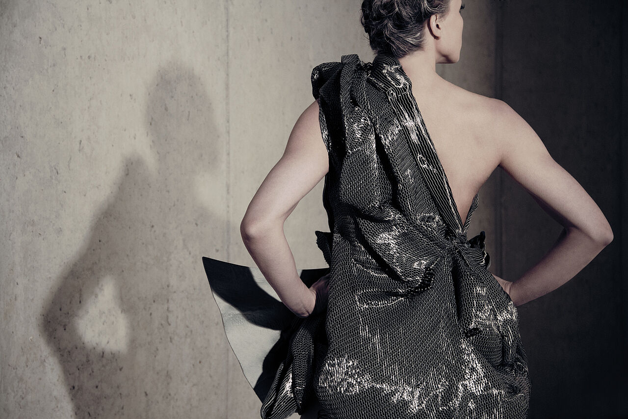 Model steht vor einer Betonwand und trägt ein Kleid aus Carbonbeton. Man sieht die Rückenansicht des Kleides.